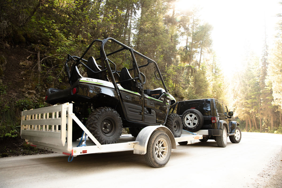 Jeep towing an open aluminum side by side trailer up a dirt road in Montana