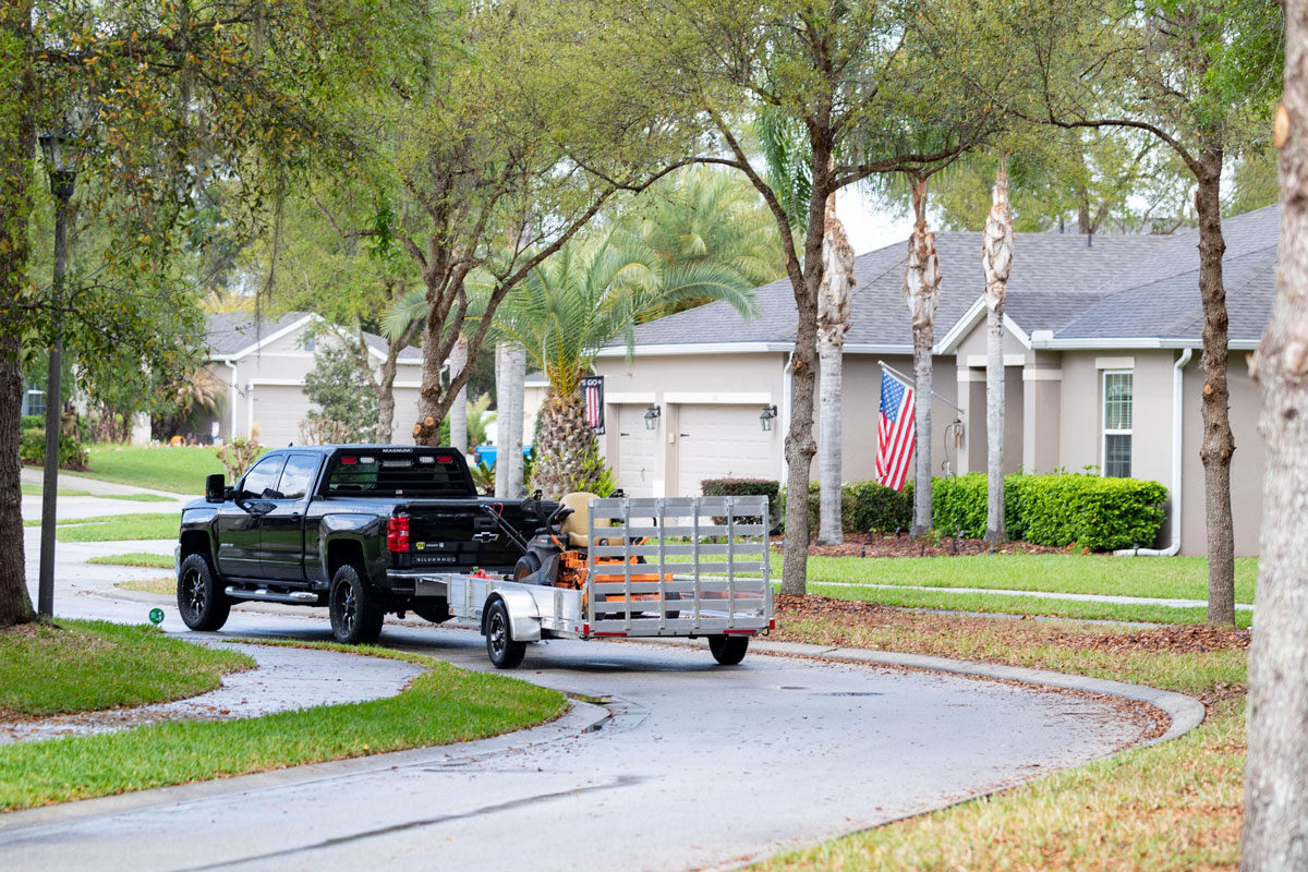 Black pickup towing AR series aluminum utility trailer with lawn mower through a neighborhood