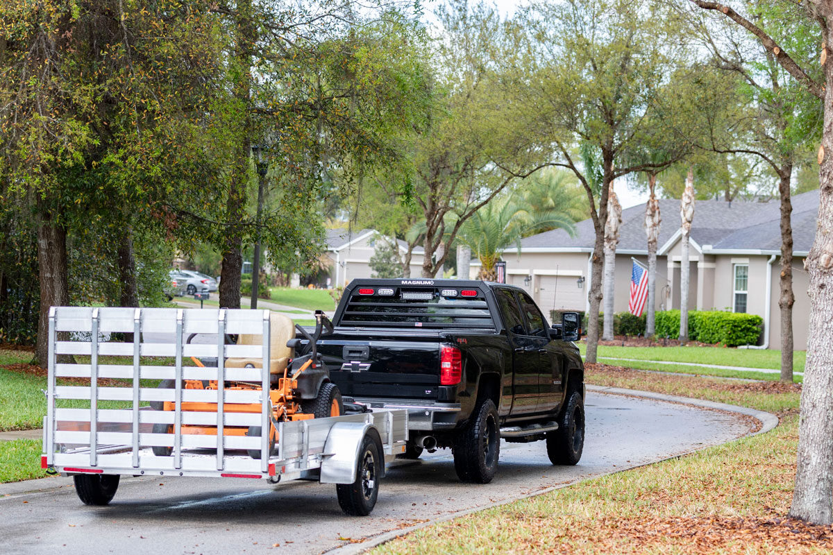 Pickup truck towing an aluminum AR trailer through a neighborhood loaded w/riding lawnmower