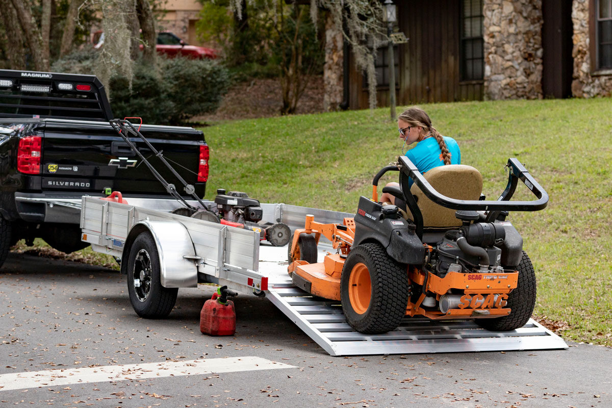 A woman unloads a riding lawnmower from the deck of an aluminum AR series utility trailer