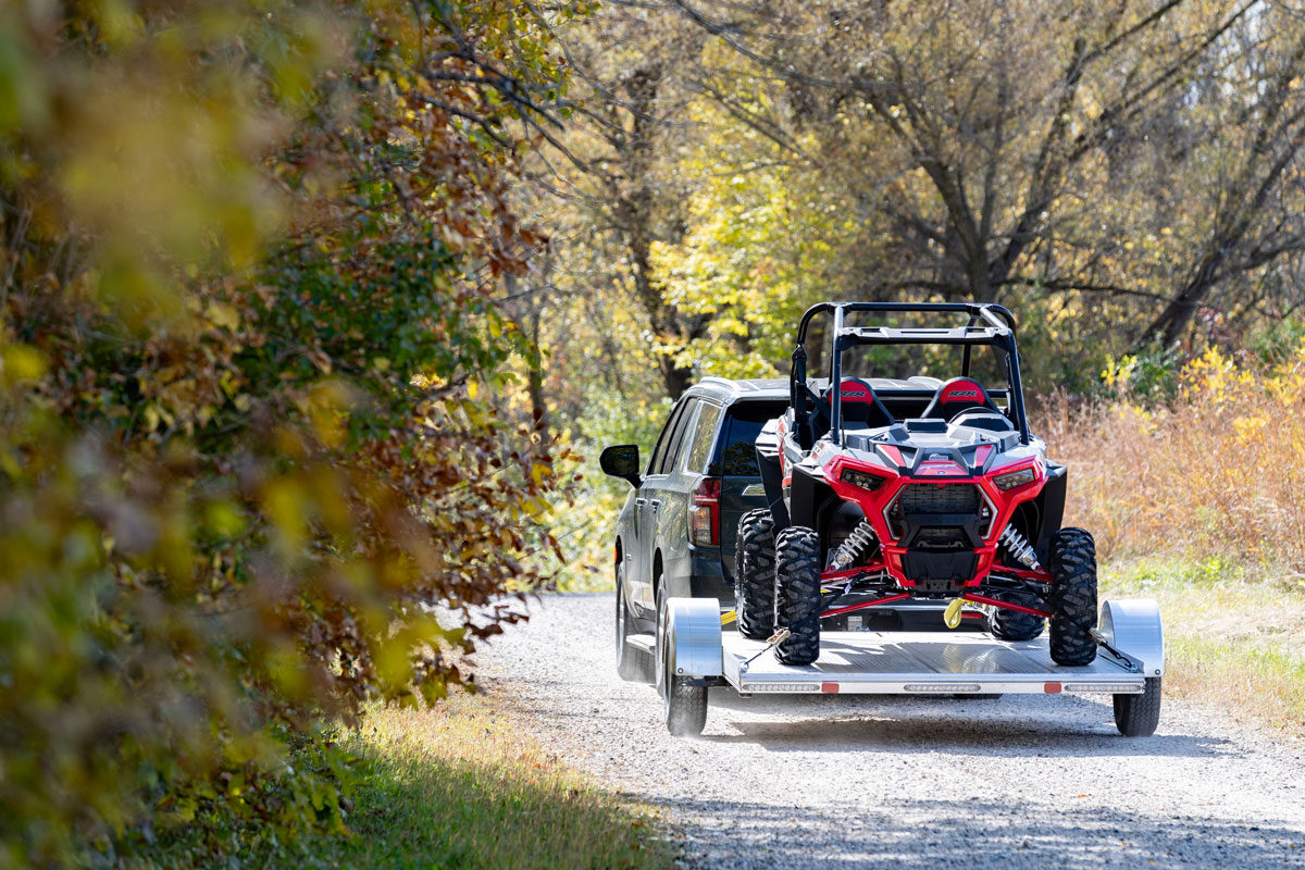 SUV towing open aluminum UTV hauler on a dirt road