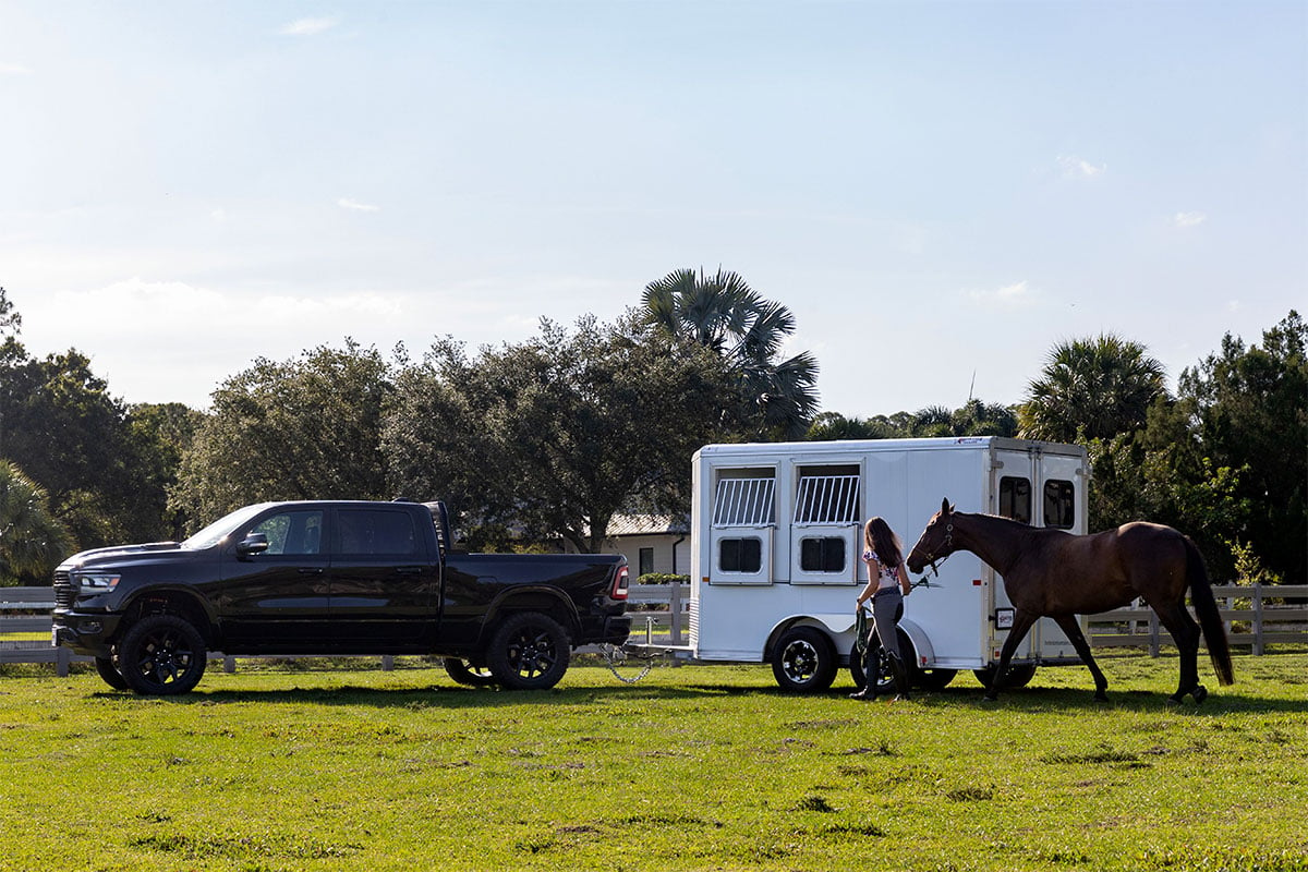 A woman leads a horse beside an aluminum Frontier horse trailer with open feed windows.