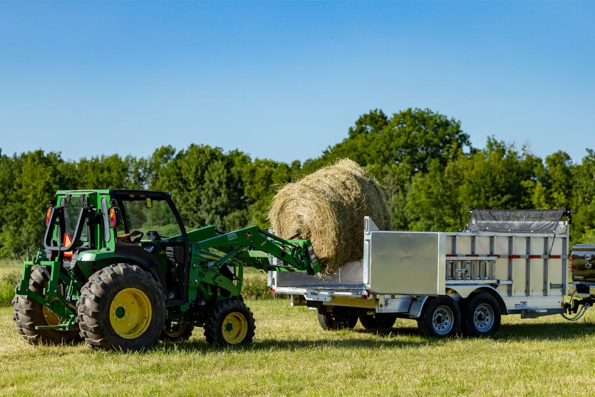 Tractor loads a large round bale of hay into a commercial aluminum dump trailer